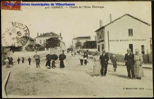 Rue du 4-Août, à la croisée avec le chemin du canal, avec de nombreux passants. Carte postale, datée par tampon de la poste du 5 octobre 1907, éd. P. Martel. [cote 2Fi10]